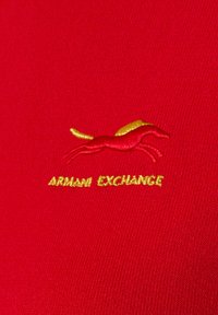 Czerwony materiał o fakturowanej powierzchni. Wzór zawiera żółte wyszywane logo konia z napisem "ARMANI EXCHANGE" poniżej, wykonanym w kolorze złotym.