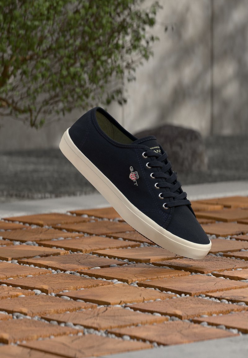 Μαύρο καμβασμένο αθλητικό παπούτσι με λευκή σόλα και κορδόνια, αιωρούμενο πάνω από ξύλινη επιφάνεια με μικρά κενά από χαλίκι, μπροστά σε τοίχο από σκυρόδεμα και πράσινο φυτό.