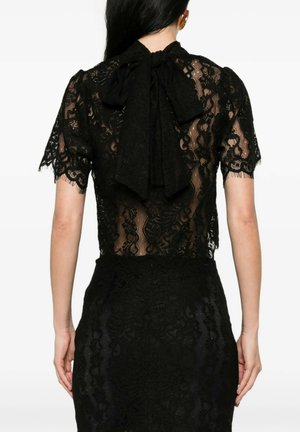 Donna che indossa un abito in pizzo nero trasparente con maniche corte e un grande fiocco legato dietro il collo, vista da dietro.