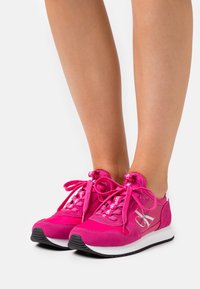 Zapatillas deportivas de ante rosa con acentos y logotipo blanco, que presentan cordones gruesos y una suela de goma texturizada. Diseñadas para comodidad y estilo.
