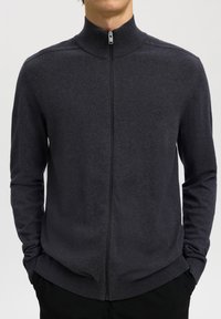 Hombre con jersey de cremallera completa de color gris oscuro con cuello alto, manos en los bolsillos, mostrado desde el cuello hasta las caderas sobre un fondo liso.