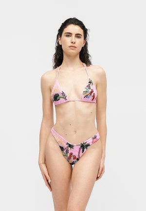 Femeie purtând un bikini roz decorat cu broderii colorate de păsări tropicale și frunziș, stând pe un fundal simplu și luminos.