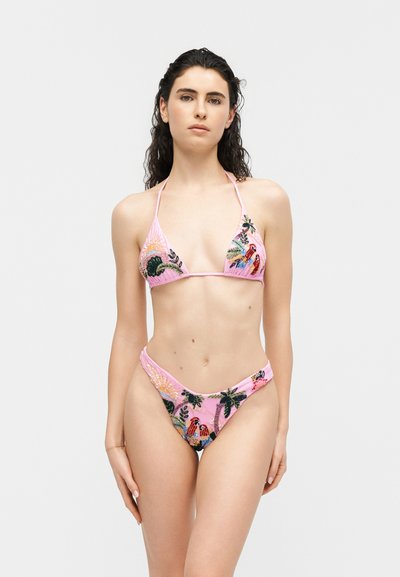 Femme portant un bikini rose orné d'oiseaux tropicaux colorés brodés et de feuillages, debout devant un fond clair uni.