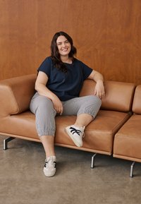 Vrouw zittend op een cognackleurige leren bank, glimlachend, gekleed in een navyblauw shirt, gestreepte broek en witte sneakers met zwarte strepen.