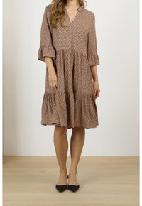 Vestido marrón con patrón de puntos, escote en V, mangas de tres cuartos en forma de campana y falda en capas. Llevado con zapatos negros de punta.