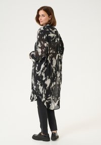 Langärmeliges, transparentes Hemd in Schwarz-Weiß mit lockerer Passform, asymmetrischem Saum und langen Ärmeln, kombiniert mit schwarzen schmalen Hosen und klobigen Schuhen.