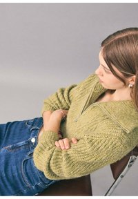 Pulover tricotat de culoare verde măslin, cu guler cu fermoar pe jumătate și țesătură texturată, asociat cu blugi din denim albastri. Modelul este așezat, cu brațele încrucișate.