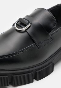 Černá kožená bota s hladkým povrchem, zdobená stříbrnou sponou a silnou, strukturální gumovou podrážkou. Design s kulatou špičkou.
