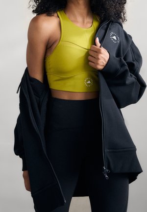 Vrouw draagt een olijfgroene sportbeha en zwarte leggings, houdt een zwarte jas met rits open met een wit logo op borst en heup.