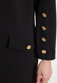 Schwarzer Stoffmantelärmel mit einer Tasche und fünf gold-texturierten Knöpfen, die entlang der Kante verlaufen. Glatte Textur und strukturiertes Design.