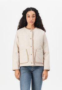 Kvadret beige jacka med rundad halsringning, tryckknappar, två framfickor och beige kant. Matchad med blå jeans.