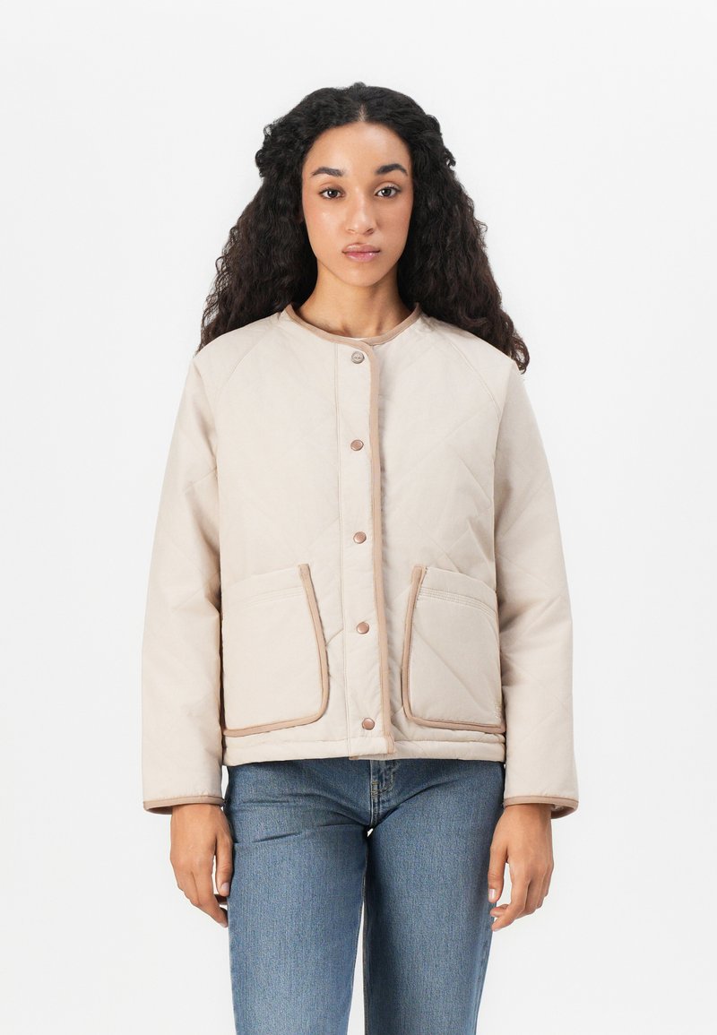 Kvadret beige jacka med rundad halsringning, tryckknappar, två framfickor och beige kant. Matchad med blå jeans.