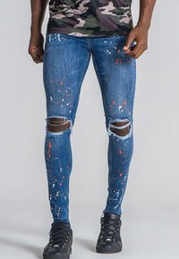 Smala denimjeans i mörkblått med slitningar vid knäna, försedd med flerfärgade färgstänk. Stylat med svarta sneakers.