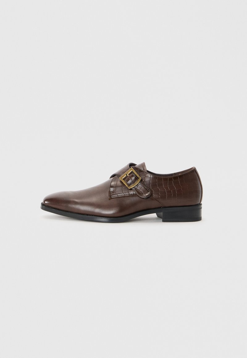 Zapato de cuero marrón con un acabado suave y patrón de cocodrilo en la correa. Presenta una hebilla dorada y un tacón negro bajo.