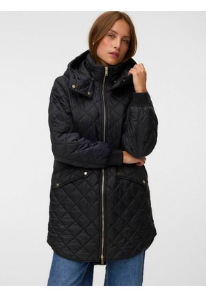 Femme portant une veste matelassée noire à capuche avec fermeture éclair avant et poches, debout les bras croisés, portant un jean bleu.