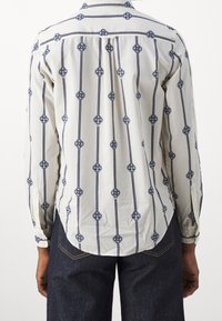 Weißes Langarmhemd mit vertikalen marineblauen Streifen und kreisförmigen Knotenmustern, getragen mit dunkelblauen Jeans, von hinten gezeigt.