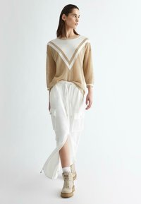 Maglione in rete beige con strisce bianche a V, abbinato a una gonna cargo bianca con fessure laterali e tasche, completato da stivali beigi in stile lace-up.