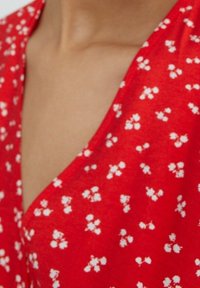 Tejido rojo con un patrón floral de pequeñas flores blancas, con un escote en V y una textura suave. La forma sigue el contorno del escote.