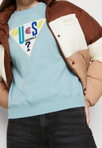 Brun och beige färgblock pufferjacka över en turkosa sweatshirt med en flerfärgad triangulär logotyp med text. Mörka jeans syns.