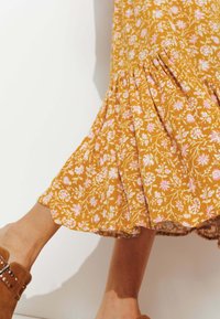 Jupe midi florale en jaune moutarde avec des motifs roses et blancs ; elle présente un ourlet évasé et des détails froncés. Des chaussures à talons avec une sangle à la cheville de couleur marron sont visibles.