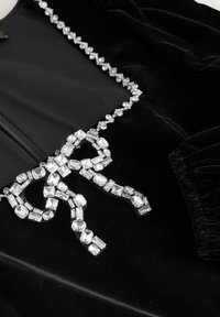 Țesătură din catifea neagră cu un design de fundă accentuată de cristale. Fundița este decorată cu cristale de diferite forme pe o lanț decorativ pentru un plus de strălucire.