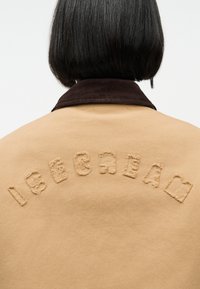 Άτομο με κοντά μαύρα μαλλιά, γυρισμένο προς τα πίσω, φοράει μπεζ μπουφάν με φθαρμένα μπεζ γράμματα που σχηματίζουν τη λέξη "ICECREAM" στην πλάτη.