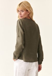 Blusa di lino verde oliva con maniche lunghe, scollatura rotonda e dettagli con bottoni sui polsini, abbinata a pantaloni di lino bianchi.