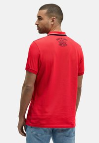 Uomo di spalle con una polo rossa con testo nero sulla parte superiore della schiena e jeans azzurri.