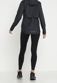 Veste légère noire avec capuche, présentant un design de dos court, associée à des leggings noirs et des chaussures de sport.