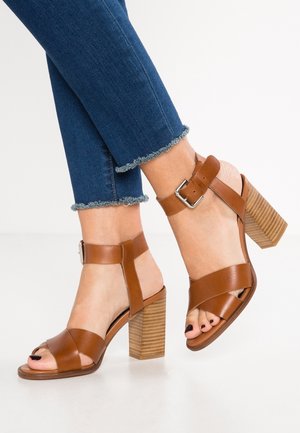 Brown leather heeled sandals with a block heel, ankle strap, and cross straps over the toes. Denim pants with frayed hem visible.