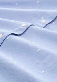 Chemise en coton bleu clair avec de petits accents brodés blancs, fermeture à boutons traditionnelle et texture lisse et douce.