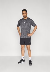 Αθλητική γκρι μπλούζα με σχέδιο tie-dye και κοντά μανίκια, συνδυασμένη με μαύρο σορτς. Λεπτομέρειες περιλαμβάνουν λευκό λογότυπο Adidas και στα δύο. Μαύρα αθλητικά παπούτσια και κάλτσες.