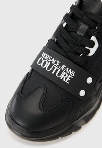 Sort sneakers med en struktureret læder- og mesh-design, tykke snørebånd, gummisål og et markant sort bånd med "VERSACE JEANS COUTURE" branding.