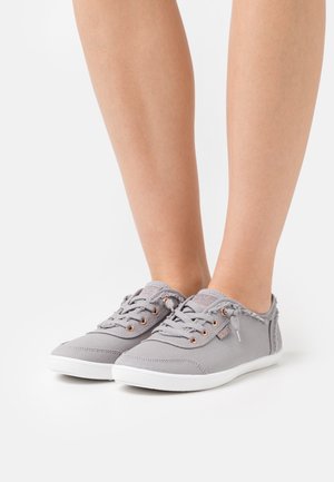 Jambes portant des baskets en toile grise à lacets avec des semelles blanches, debout sur une surface blanche. Les chaussures présentent des bords effilochés le long du col.