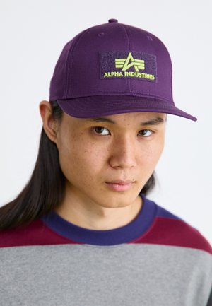 Purple baseball cap with a textured black patch featuring the "Alpha Industries" logo in yellow. Cotton material. Adjustable at the back.