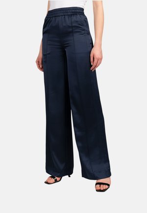 Donna che indossa pantaloni in raso blu navy a vita alta e gamba larga e sandali neri con tacco e punta aperta, in piedi davanti a uno sfondo semplice.
