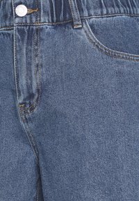 Džínové kalhoty s tmavým praním, které mají elastický pas, oranžové prošívání a kapsy. Materiál má hladkou strukturu s mírným leskem.