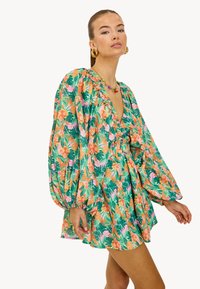 Mujer vistiendo un vestido corto de flores con estampado tropical en verde, naranja y rosa, con mangas largas de globo y un escote en V profundo.