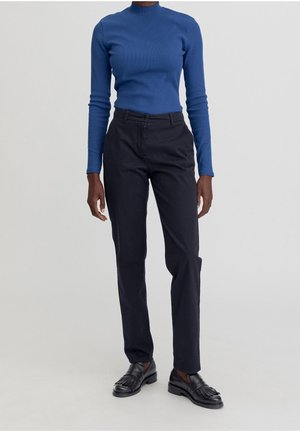 Osoba ubrana w niebieską bluzkę z długim rękawem, czarne spodnie na miarę oraz czarne skórzane mokasyny z frędzlami, stojąca na jednolitym tle.