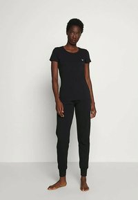 Sieviete stāv basām kājām, tērpusies pieguļošā melnā īsajās piedurknēs krekliņā un melnās sporta biksēs, pret vienkāršu baltu fona.