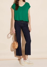 Grön kortärmad topp, mörk denim raka jeans, krämfärgade sneakers med rosa detaljer, och en rund vävd väska med axelrem.