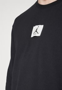 Czarny sweter z okrągłym dekoltem, z białą łatą z logo i podpisem w czerni. Miękka tekstura, prosta forma.