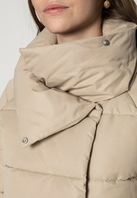 Beżowa kurtka puchowa z dużym, złożonym kołnierzem, detalem na zatrzask, pikowaną teksturą i gładkim materiałem. Zbliżenie na obszar dekoltu.