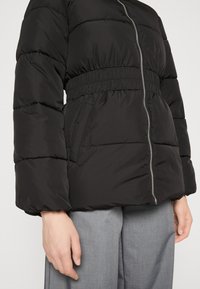 Crni puffer kaput s elastičnom pasicom, srebrnim zatvaračem i dva bočna džepa. Ima oversized rukave s glatkom, mat površinom.