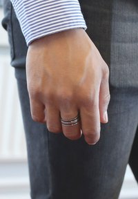 Zilveren ringen gestapeld op een hand, die een blauw-wit gestreept shirt draagt. De hand is dicht bij een grijze broek met een gestructureerd oppervlak gepositioneerd.