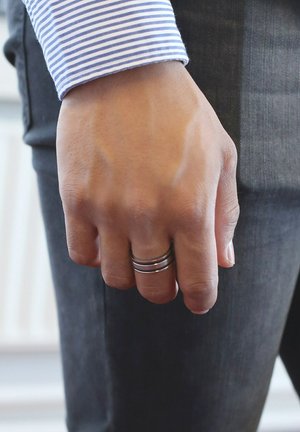 Des bagues en argent empilées sur une main, portant une chemise à rayures bleues et blanches. La main est positionnée près d'un pantalon gris avec une surface texturée.