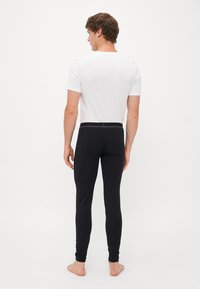Man som står barfota med ryggen mot kameran, klädd i en vit t-shirt med kort ärm och svarta åtsittande leggings mot en enfärgad vit bakgrund.