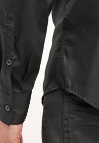Camicia nera lucida con un design elegante. Presenta un bottone visibile sul maniche e un tessuto liscio e testurizzato. Vestibilità su misura.