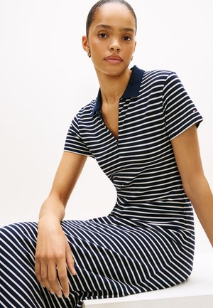 Γυναίκα με πίσω μαλλιά που φοράει φόρεμα πόλο με ναυτικό μπλε και λευκές ρίγες, καθιστή, κοιτάζοντας μπροστά με ουδέτερη έκφραση.