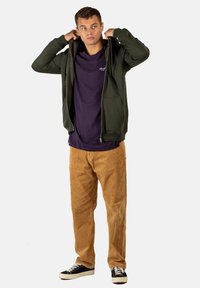 Sudadera verde con cremallera sobre una camiseta morada oscura, combinada con pantalones de pana color beige y zapatillas negras. Tela suave y corte casual.